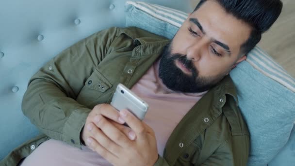 Bonito mestiço usando smartphone desfrutando de mídia social relaxante no sofá em casa — Vídeo de Stock