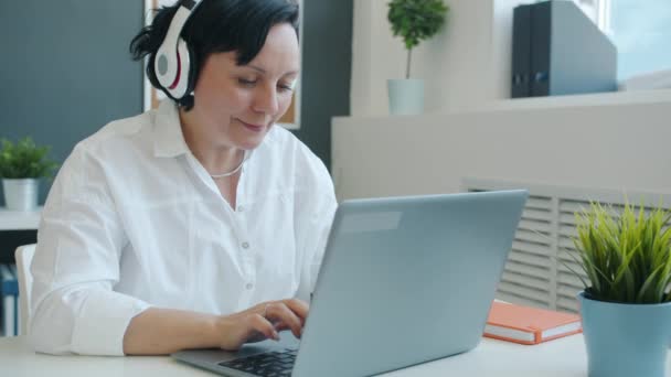 Радостная деловая женщина, работающая с ноутбуком и наслаждающаяся музыкой через наушники в офисе — стоковое видео