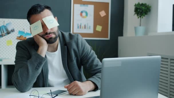 Zmęczony biznesmen drzemka w biurze siedzi przy biurku mając lepkie notatki z otwartymi oczami zdjęcia zakrywające twarz — Wideo stockowe