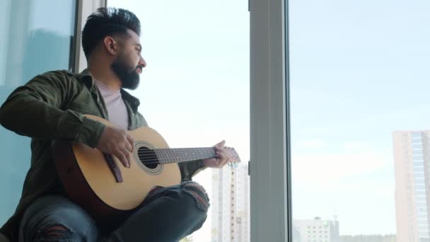 Повільний рух молодого близькосхідного чоловіка, який грає на гітарі, сидячи на підвіконні вдома. — стокове відео
