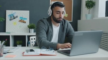 Keyifli bir ofis çalışanı dizüstü bilgisayar kullanıyor ve kulaklıkla müzik dinliyor sonra da masada dans ediyor.