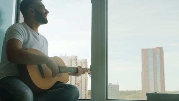 Vrolijke bebaarde man gitarist spelen muziekinstrument en zingen dan lachen zittend op vensterbank — Stockvideo