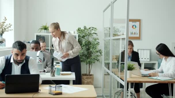 Radosna kobieta rozmawia z kolegą otrzymując dobre wieści rzucając papierami i tańcząc w biurze — Wideo stockowe