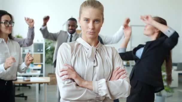 Poważna bizneswoman z blond włosami stojąca w biurze podczas gdy współpracownicy tańczą w tle — Wideo stockowe