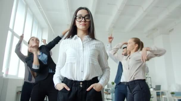 Joven mujer de negocios asiática con cara seria de pie en la oficina donde las personas hombres y mujeres bailan — Vídeos de Stock