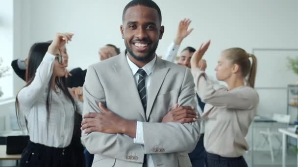 Empresario afro-americano confiado parado en oficina sonriendo mientras que la gente que baila en fondo — Vídeo de stock