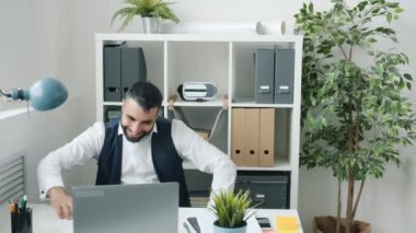 Neşeli girişimci laptopla çalışıyor sonra da iş arkadaşlarıyla dans edip başarıyı kutluyor.