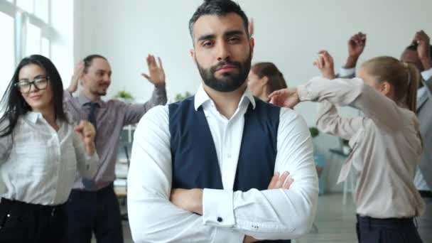 Biznesmen w średnim wieku stoi w biurze z poważną twarzą podczas gdy ludzie tańczą świętując osiągnięcia biznesowe — Wideo stockowe