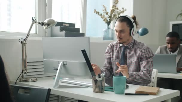Kreativer junger Mann mit Kopfhörern tanzt mit Computer am Schreibtisch im Büro — Stockvideo