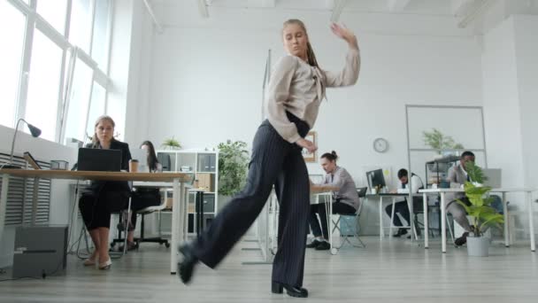 Красивая молодая женщина исполняет современный творческий танец на рабочем месте, в то время как коллеги заняты работой — стоковое видео