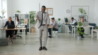 Takım elbiseli insanlar arka planda çalışırken modern ofiste dans eden Afro-Amerikalı sanatçı.