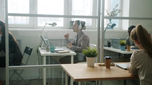 Alegre chico usando auriculares bailando relajante en el lugar de trabajo mientras la gente ocupada trabajando — Vídeos de Stock
