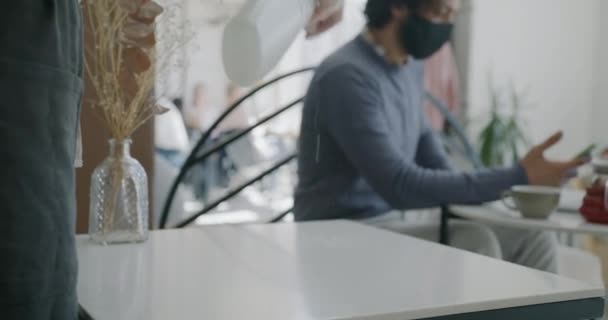 Близко к официантке дезинфицирующий стол в то время как парень клиент носить маску едят на заднем плане — стоковое видео