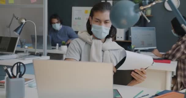 Άνδρας υπάλληλος που φοράει ιατρική μάσκα και εργάζεται στο γραφείο χρησιμοποιώντας φορητό υπολογιστή και ελέγχοντας έγγραφα — Αρχείο Βίντεο