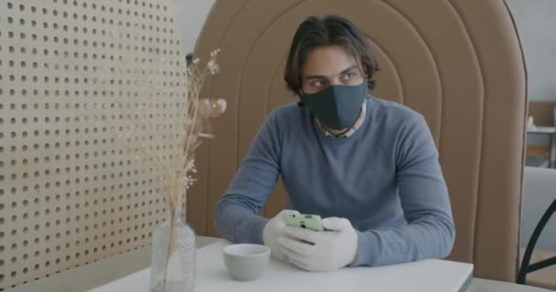Jeune homme arabe portant un masque à l'aide d'un smartphone à la table du café lorsque la serveuse apporte de la nourriture et des boissons — Video