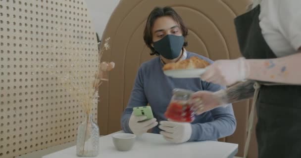 Bliskowschodni facet w masce i rękawiczkach za pomocą smartfona i rozmowy z kelnerką w kawiarni — Wideo stockowe