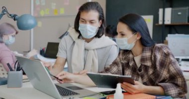 Erkek ve kadın meslektaşlar laptopla çalışıyor ve ofiste yüz maskeleri takarak konuşuyorlar.