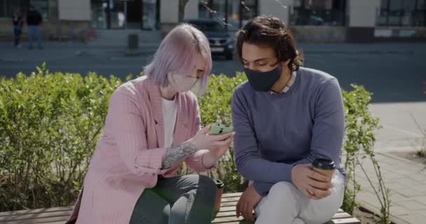 Chica y chico usando smartphone bebiendo para ir a tomar café hablando y riendo afuera durante la pandemia — Vídeo de stock