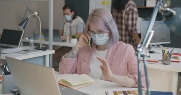 在大流行病期间，戴着医疗面罩的年轻女士在办公室里用手机交谈 — 图库视频影像