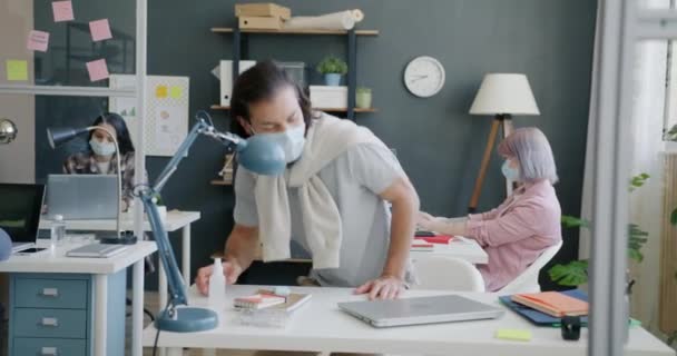 Giovane uomo che indossa maschera venire in ufficio saluto colleghi spruzzando le mani con disinfettante — Video Stock