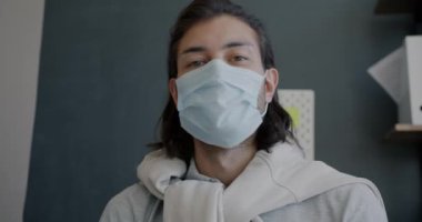 Yüz maskesi takan bir adamın portresi ofiste görüntülü konuşma yapıyor ve kameraya bakıyor.