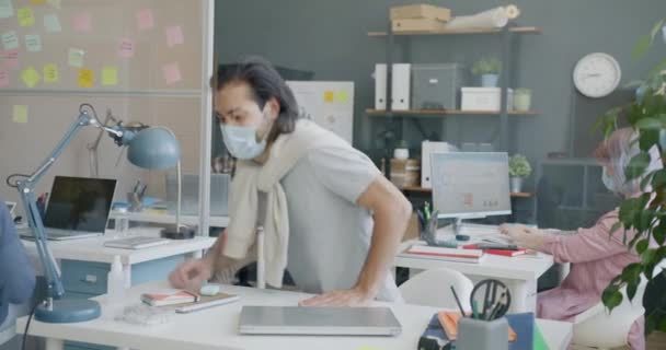 Pracownik w masce przychodzi do pracy powitanie kolegów dezynfekujących ręce i za pomocą laptopa — Wideo stockowe