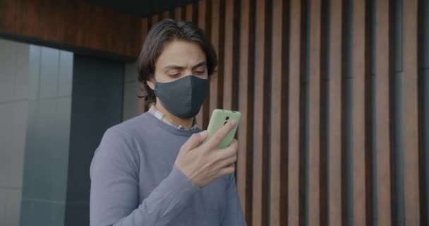 Movimento lento di attraente ragazzo del Medio Oriente che indossa maschera facciale utilizzando smartphone e camminare fuori — Video Stock