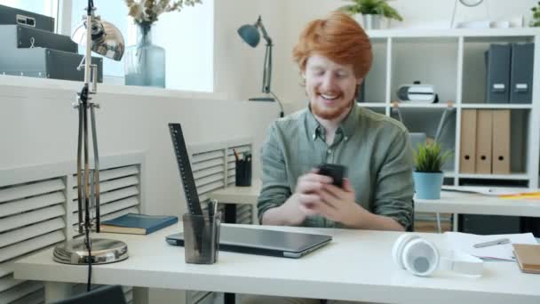 Радісний рудий чоловік вказує на смартфон і посміхається, сидячи за столом в офісі, дивлячись на камеру — стокове відео