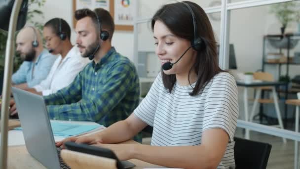 Fröhliche junge Callcenter-Mitarbeiterin spricht und tippt mit modernen Geräten mit Kollegen im Hintergrund — Stockvideo