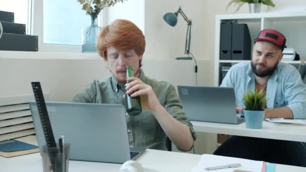 П'яний чоловік тримає пляшку пива і п'є, працюючи з ноутбуком, а потім спить, коли колеги звинувачують — стокове відео