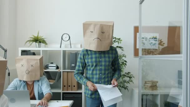 办公室职员，头戴纸袋，在办公桌前工作时表现得情绪低落，而脾气暴躁的老板则给他们写文章 — 图库视频影像
