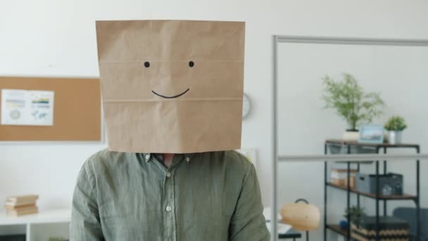 Portrait of male employee wearing paper bag with smile emoji standing in office and looking at camera — Stock Video