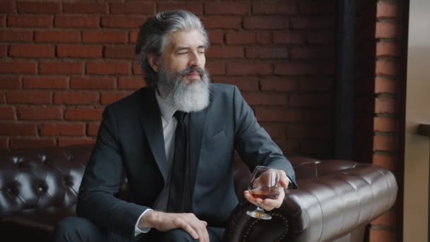 Grå-hårig man i elegant kostym sitter på läder soffa hemma håller glas konjak — Stockvideo