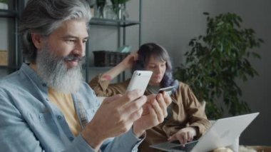 Akıllı telefon uygulaması üzerinden alışveriş yapan heyecanlı adam elinde kredi kartıyla karısının onaylamadığı ekranı göstererek gülüyor.