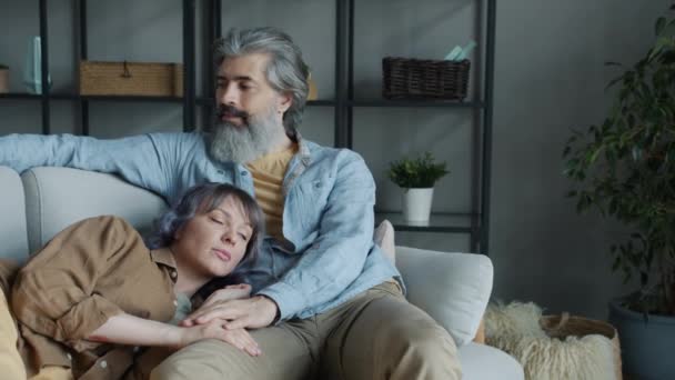 Hombre maduro usando teléfono inteligente y abrazando a la hermosa esposa acostada en su regazo en casa — Vídeos de Stock