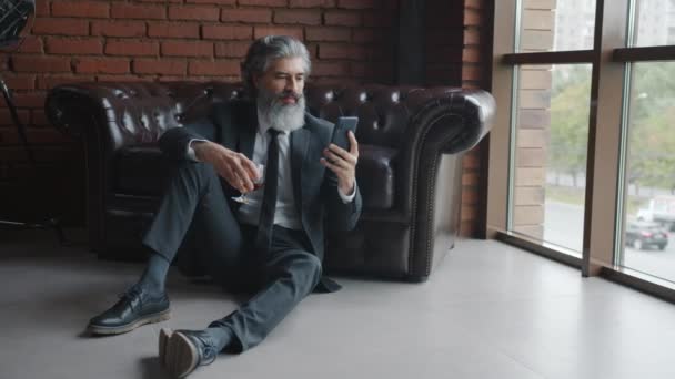 Zpomalený pohyb pohledný dospělý muž pomocí chytrého telefonu a drží sklenici koňaku sedí na podlaze doma — Stock video