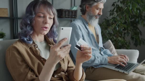 Mouvement lent de femme joyeuse faisant du shopping en ligne tout en mari malheureux en utilisant un ordinateur portable et secouant la tête à la maison — Video