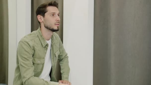 Retrato de un tipo infeliz gruñendo esperando a una mujer que está probando ropa en el probador — Vídeos de Stock