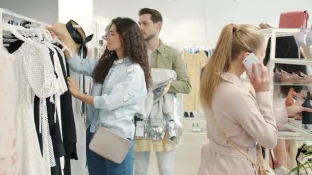 Młoda kobieta robi zakupy z mężem wybierając ubrania, podczas gdy wkurzony mężczyzna wyraża negatywne emocje — Wideo stockowe