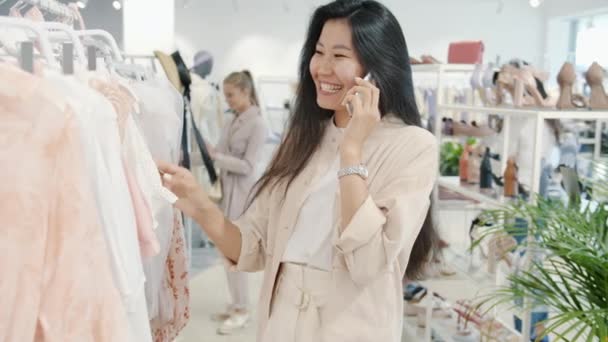 Mulher asiática atraente conversando no telefone celular e olhando para roupas na loja de moda — Vídeo de Stock