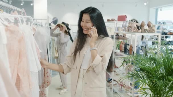 Câmera lenta de feliz senhora asiática escolhendo roupas na loja e falando no telefone móvel — Vídeo de Stock