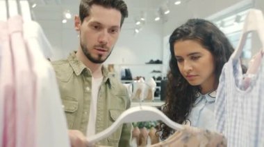 Neşeli çift giysi seçiyor. Kıyafetleri tartışıyor. Modern mağazada askıdaki gömleklere dokunuyor.