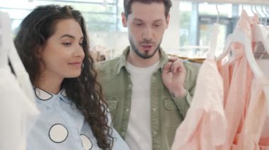 Kadın dükkanda kıyafet seçerken, erkek fiyatlardan memnun değil.