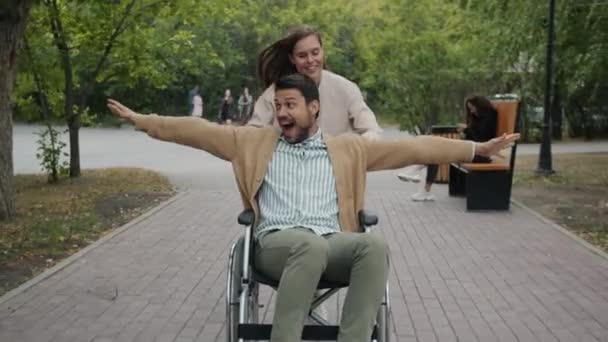 障害者の男は公園で笑顔で速い乗り心地を楽しんでいる間、女性のドリーショットを実行し、車椅子を押す — ストック動画