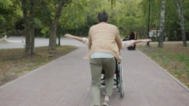 Bakıcı koca koşuyor ve tekerlekli sandalye itiyor. Engelli karısıyla sonbahar parkında eğleniyor.