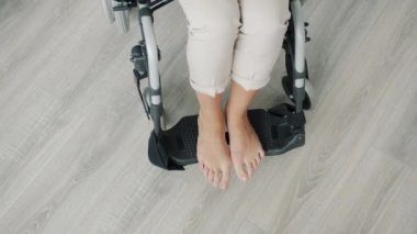 Evdeki modern tekerlekli sandalyede çıplak kadın ayaklarının yüksek açılı görüntüsü.