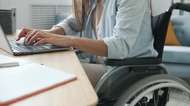 Nierozpoznawalna niepełnosprawna dziewczyna korzystająca z laptopa, pisząca na laptopie, siedząca na wózku inwalidzkim przy biurku zajęta pracą online — Wideo stockowe