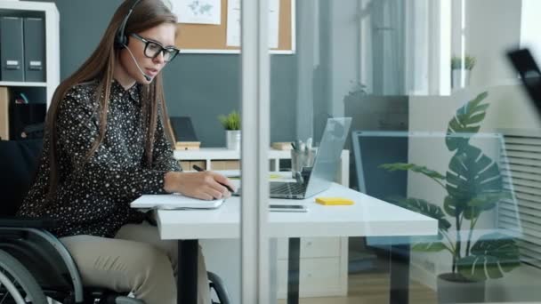 Wesoły niepełnosprawna dziewczyna pracownik rozmawia z klientami online z laptopem i słuchawki śmiejąc — Wideo stockowe