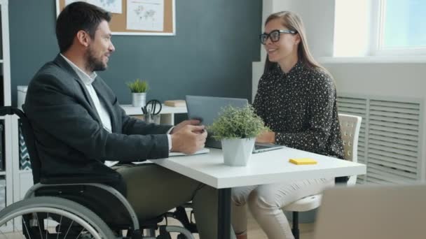 Gehandicapte man in een rolstoel die zaken bespreekt met vrouwelijke collega die aan zijn bureau zit te praten — Stockvideo