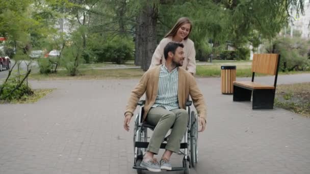 Liebevolle Frau schiebt Rollstuhl mit fröhlichem Mann beim Spazierengehen im Herbstpark — Stockvideo